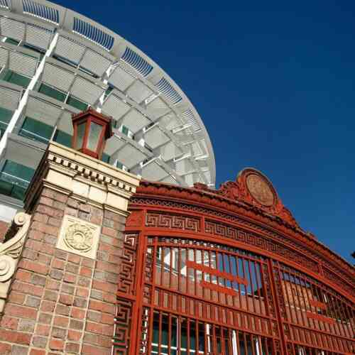 A white circular building with many windows is seen with a red gate and brick pillars in front of it. It is a sunny day with blue skies