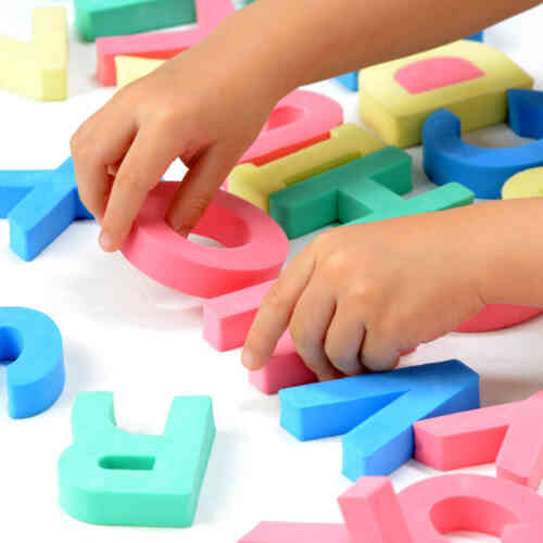 A child with light skin is seen touching colorful letters with their hands as they try to spell something out. The letters are green, blue, red, and yellow.