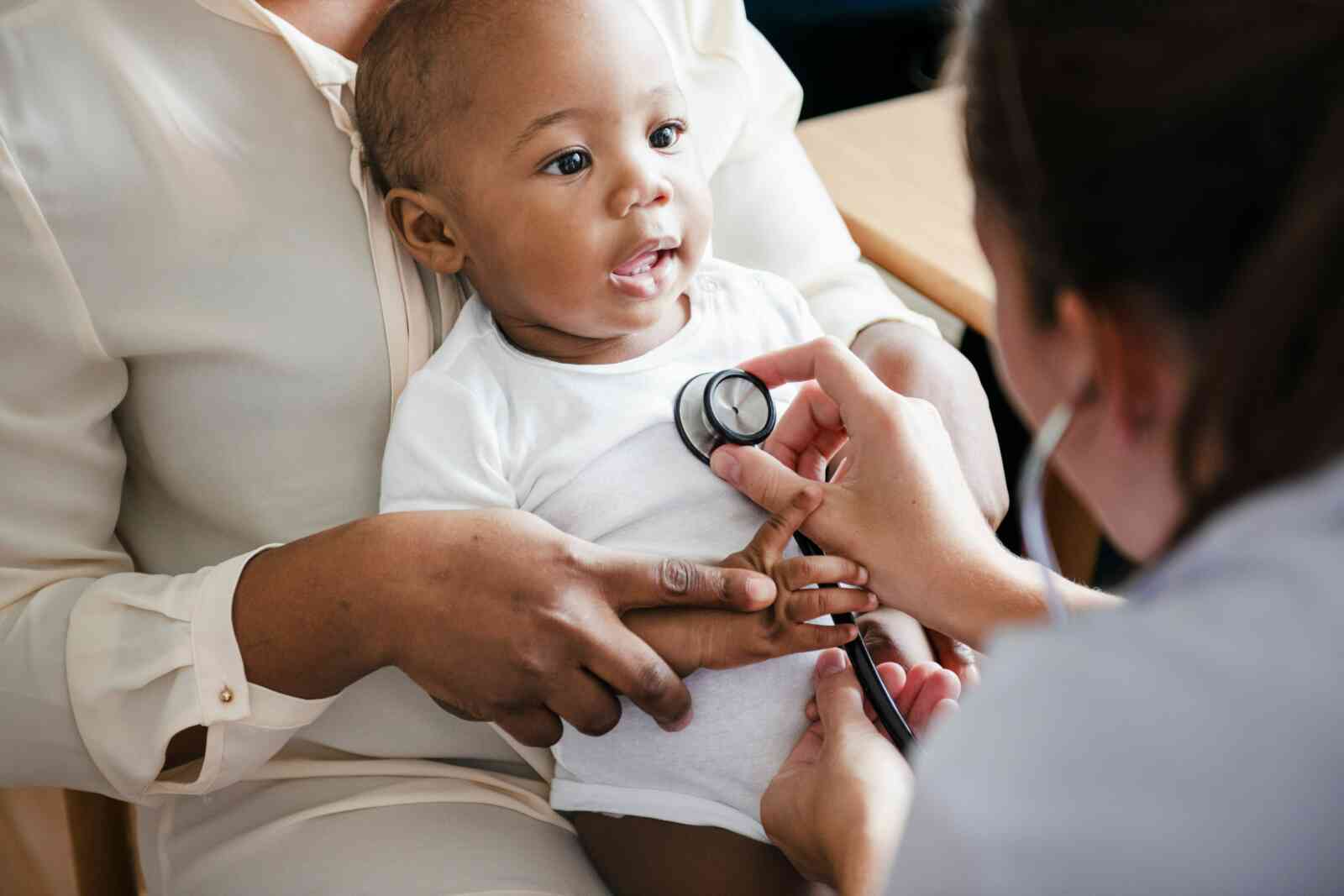 A baby is sitting on the lap of an adult individual whose face cannot be seen but their hand is placed on the baby's arm. The baby is facing an adult individual who appears to be a doctor and has a stethoscope placed on the baby's chest. The baby is smiling. The baby and adult they are sitting on have dark brown skin and the other individual has light skin.