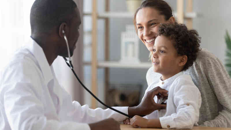 An adult female is standing behind a young child smiling while the child smiles at a doctor who is listening to the child's heart with a stethoscope. the adult female and child have light brown skin and the doctor has dark brown skin.