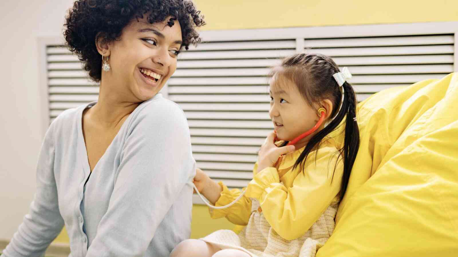 An adult female is sitting next to a young child who is sitting on a bright yellow bean bag chair. The young child has a toy stethoscope in her ears and has the other end placed on the adult female's back. They are smiling at one another. Both individuals have light brown skin.