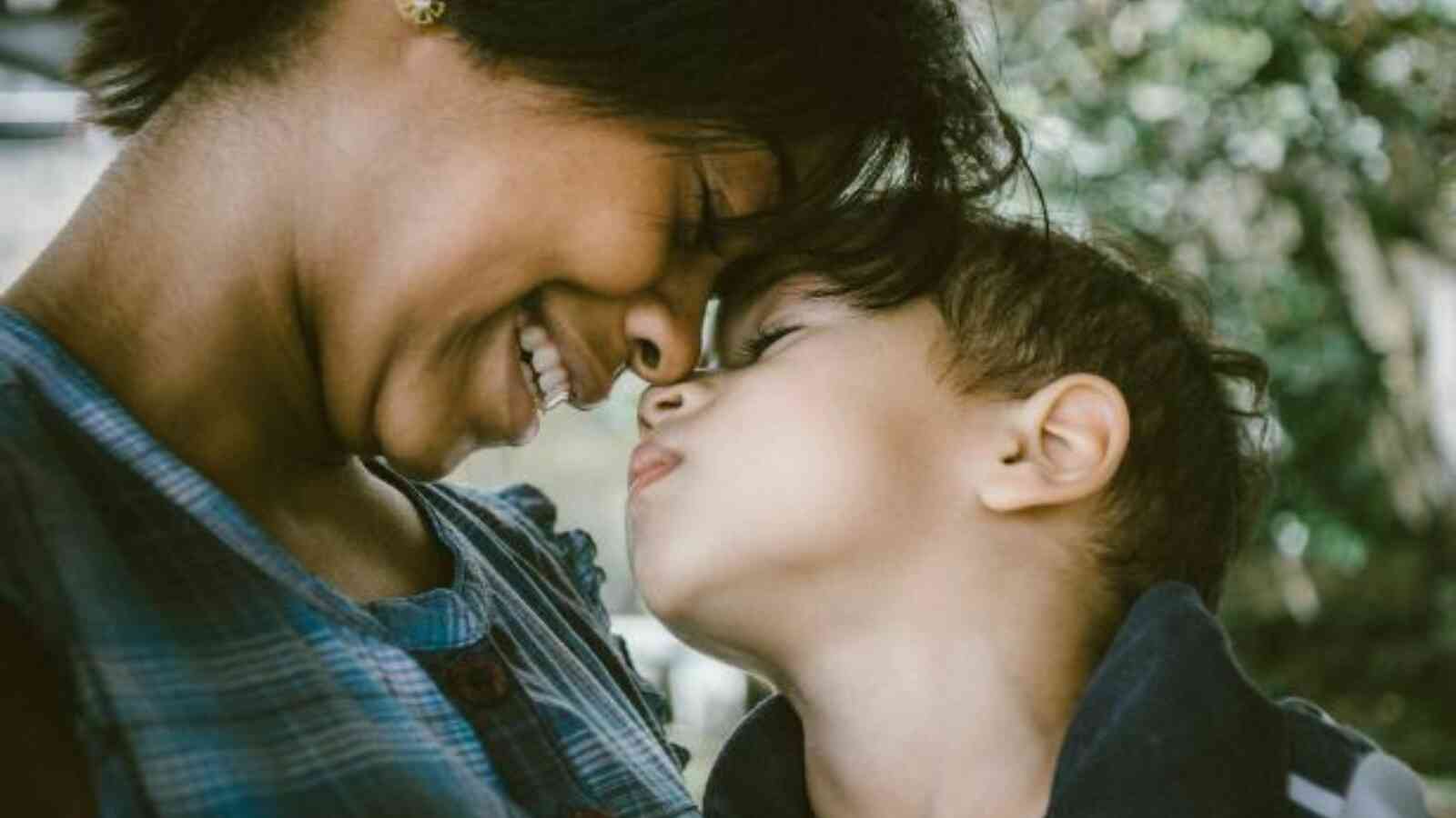 A young child and an adult female who appears to be the caregiver, are leaning into one another and touching noses. The adult female is smiling and both individuals have their eyes closed. The adult female has dark brown skin and the child has light brown skin.