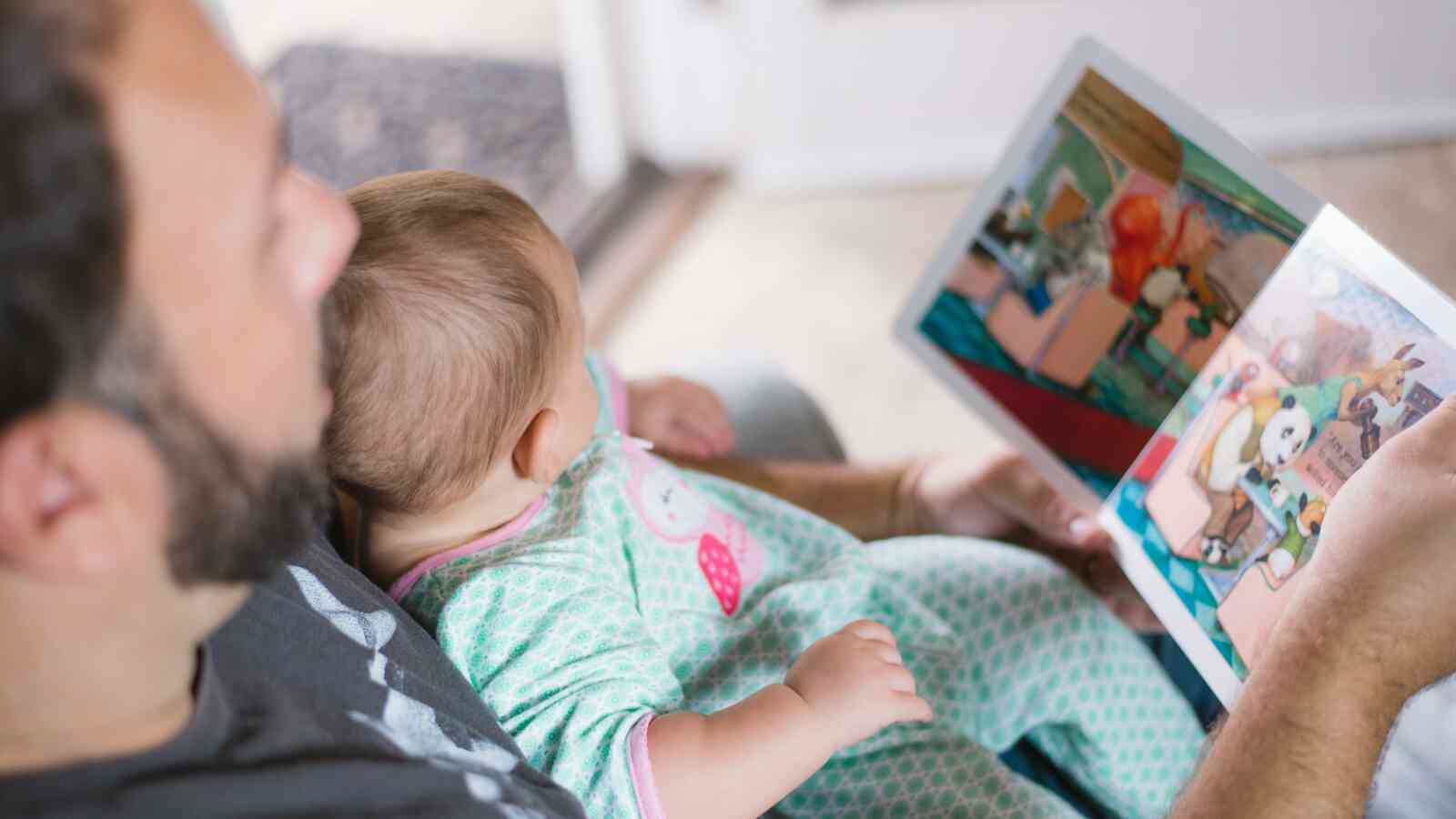 An adult male is seen sitting with a baby on his lap. The baby and adult male are looking at a book that he has open with colorful pictures. Both individuals have light skin.