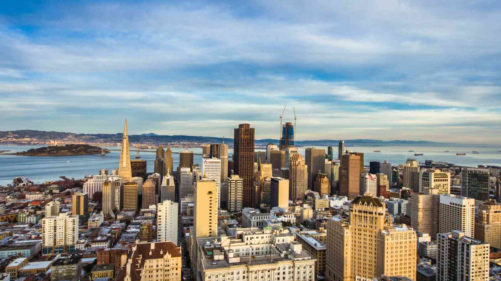 A skyline of the city of San Francisco on a sunny day with blue skies and some clouds. There are tall buildings in the center of the image and water from the bay can be seen in the distance.